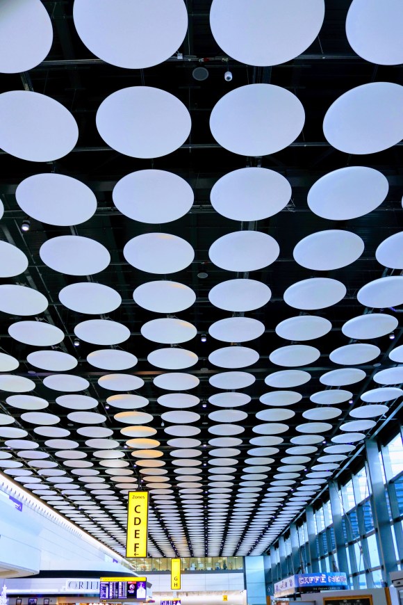 a concourse at Heathrow airport with a spotted ceiling of white sound proofing discs
