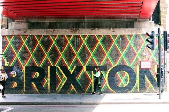 a painted 'BRIXTON' mural  with geometric green, yellow and red hatched stripes on a wall under the main bridge by the station