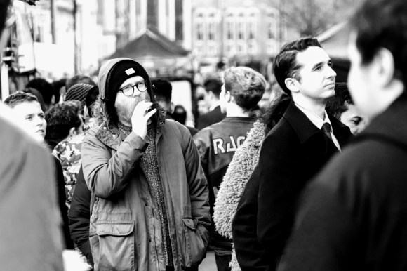 Lunch time food shoppers browing what to chose at a pop-up food market. The main character is focussing inteently on the menu: he's wearing a wolly hat and parka typw hoodie, and vaping