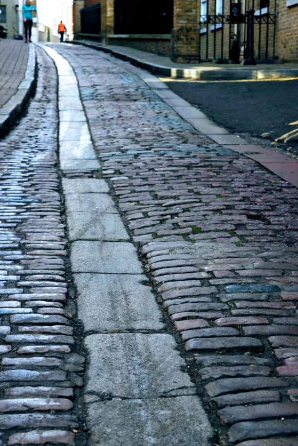 looking up at a grey cobbled street with two distant pedestrians in the background providing a blue and orange colour contrast