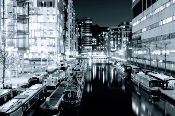 a night time view of the Thames Basin with high rise offices on either side of the water and their lights and those from moored barges reflect on the still water