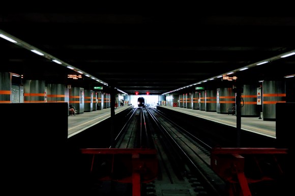 a dark picture with a train emerging into the station backlit by the sunlight outside. Pillars on either side of the platfporm draw your view towards the train