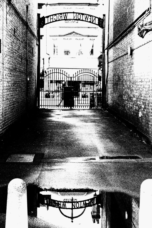 an old works gated entrance for 'Newton Wright Limited' taken in black and white and a reflection captured in a still puddle in the foreground