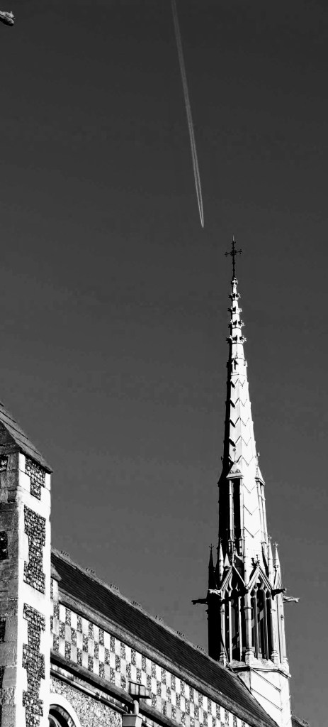 an overhaed aeroplane with its vapour trail appearing to almost colide with the tip of the church steeple and cross