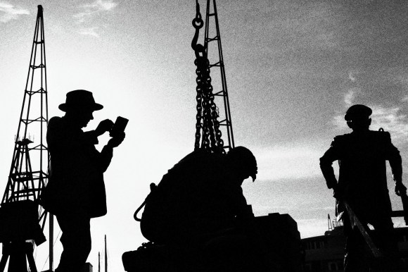 a black and white image os a statue of three dockers going about their business. The background also captures the shadowy image of some of the cranes used in the docking heyday