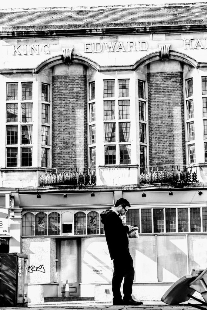 A black and white phot of a man standing still looking at his phone opposite King Edward Hall along Finchley Road