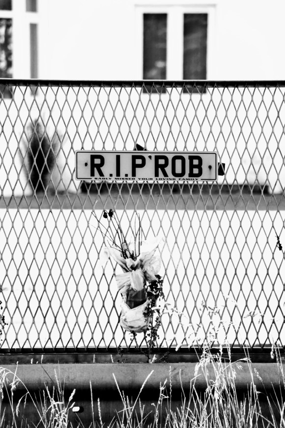 A black and white photo of a car number plate style RIP Rob sign attached to the dual carriageway central reservation barrier with a decayed bouquet of flowers