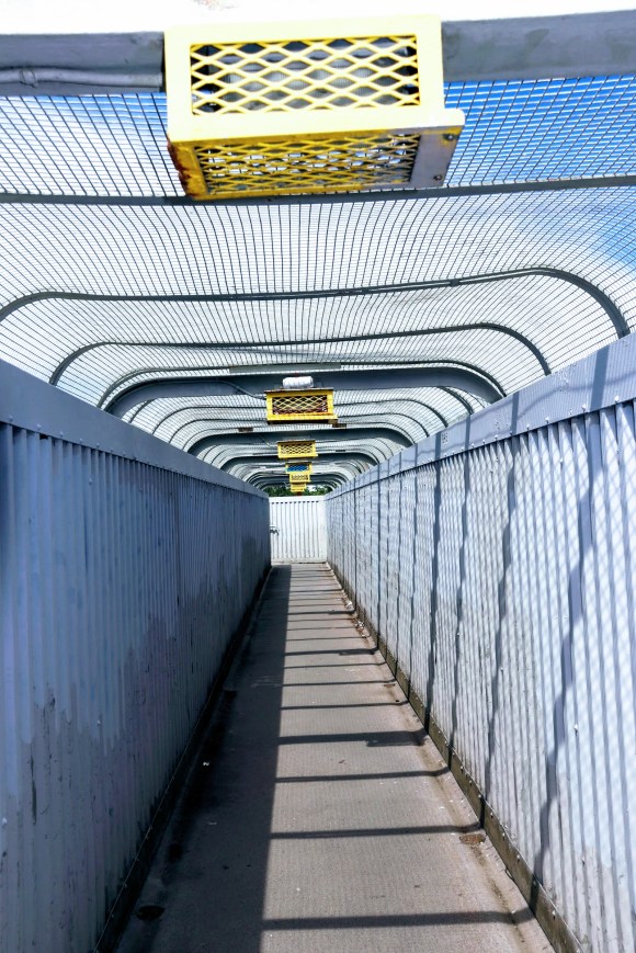 A colour photo looking along the length of a footbridge with tall sides and a mesh canopy. In the bright sunlight, the blue/grey metalwork contrasts with the yellow overhead light guards