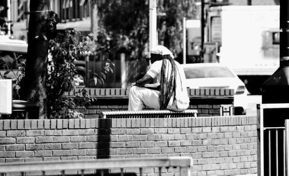 A black and white photo of a gent dressed in white shirt, trousres and headcap covering long dreadlocks who's sitting on a street bench