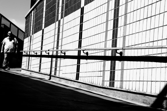 A black and white photo with diagonal lines of this shadowy walkway edging from middle left to bottom right. A gent walking into shot at middle left helps to balance the picture
