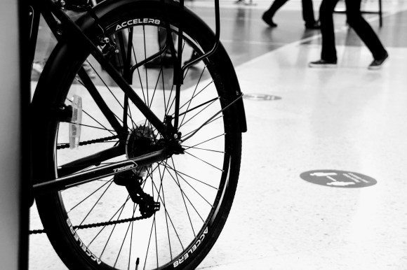 a blck and white shot of a bicycle tyre in the foregoround emblazoned with the makers name 'Acceler8'. Passengers feet walking by in the background