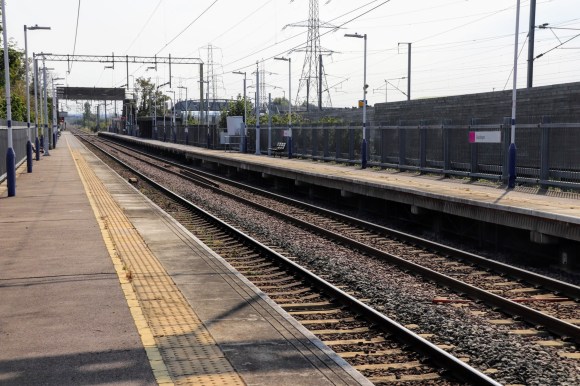 a lomg view along the length of an empty platform with the track extending as far as the eye can see. Overhead cables and nearby electricity pylons dominate the skyline