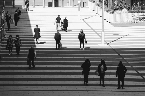Pedestrians walking along the side of the main station. The walkway has a strupped light and dark tiled layout