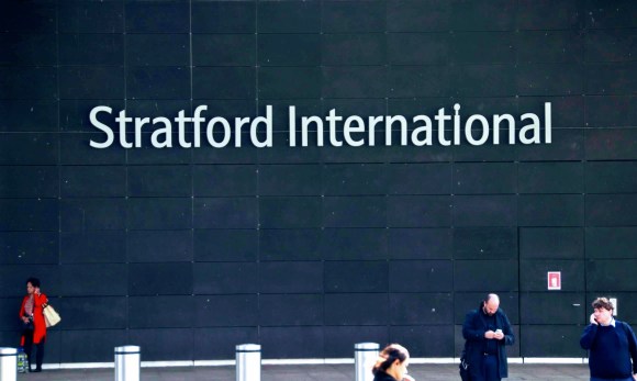 'Stratford International' sign on the side of the sttaion with four people in the foreground. One lady in a red coat stands out as she talks on her mobile.