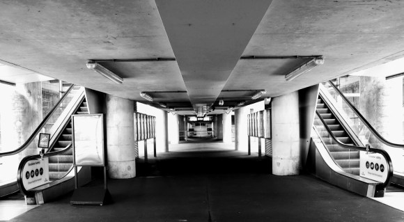 tunnel view to a waiting room in the centre of the picture, flanked on each side by pillars, notice boards and an escalator