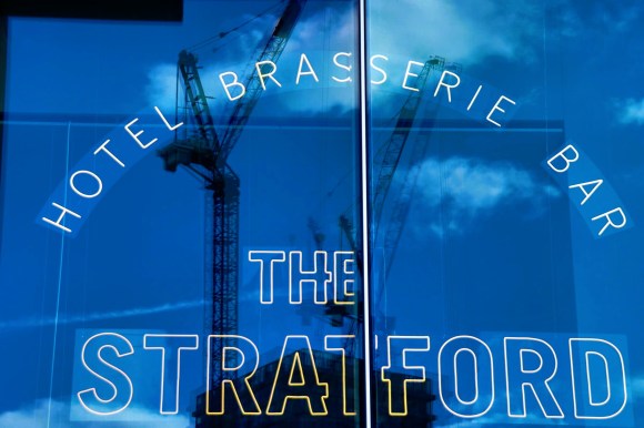 The entrance to 'The Straford', outline letters on the bottom of the picture and above in an arc 'Hotel Btasserie Bar'. The windown reflecting a blue sky and two cranes reflected