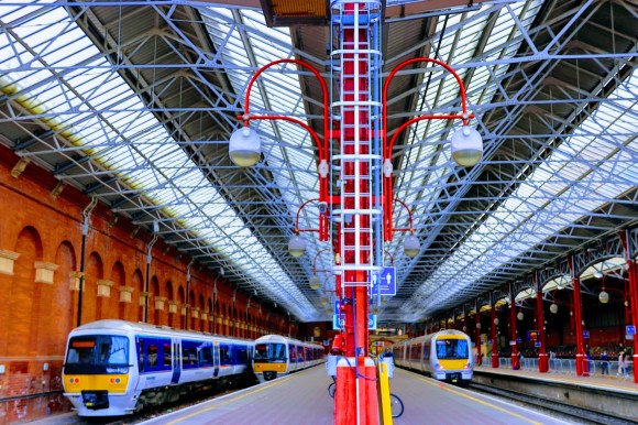 an enhanced colour picture standing of platforms 1 and 2 with three rolling stock waiting to depart. The colour scheme is predominantly red on the supporting roof pillars, blue on the roof support structure and brick on the supporting sttaion wall