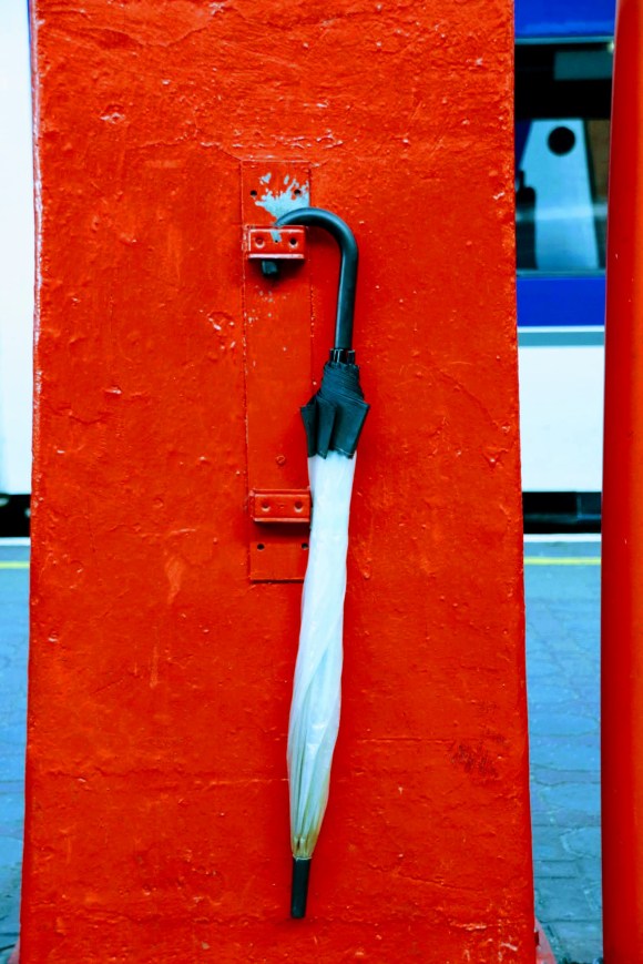 a white umbrella with black fringes hanging, rolled up, on a hook fixed to one of the red roof support pillars along the length of platforms 1 and 2