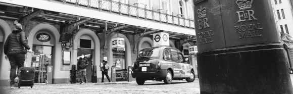 a black and white image of the station's forecourt with EIIR post box on right hand side, taxi  right centre and  a pedestrian making their way into the station pulling a wheelie suitcase