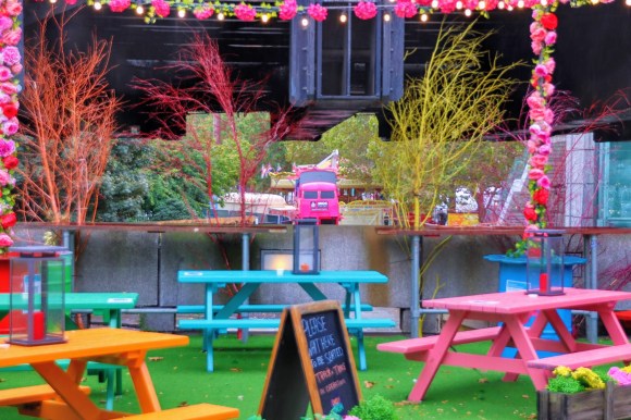 colour photo. The centrepiece in the distance is a pink 'Snog' double decker bus. The bus is frames through the colourful furniture display of the open air Beany Green eaterie. Picnic tables in green, orange, pink and cyan, framed by a string of pink flowers