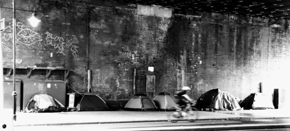 black and white image under Hungerford bridge of 6 camping tents of different sizes up against the bridge wall. A blurred passing cyclist gives a slight reference to speed