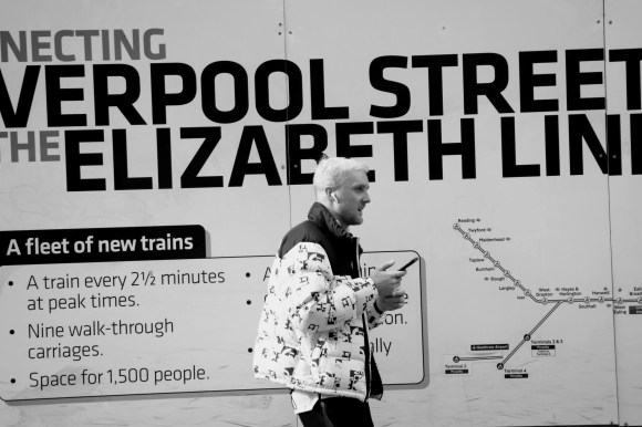 Black & White: building hoarding disaplaying part o 'connecting Liverpool Street - The Elizabeth Line. Standing in front is a blond haired young gent talking on his mobile phone looking to the right. He's wearing a predominantly white puffer jacket with black lettering.