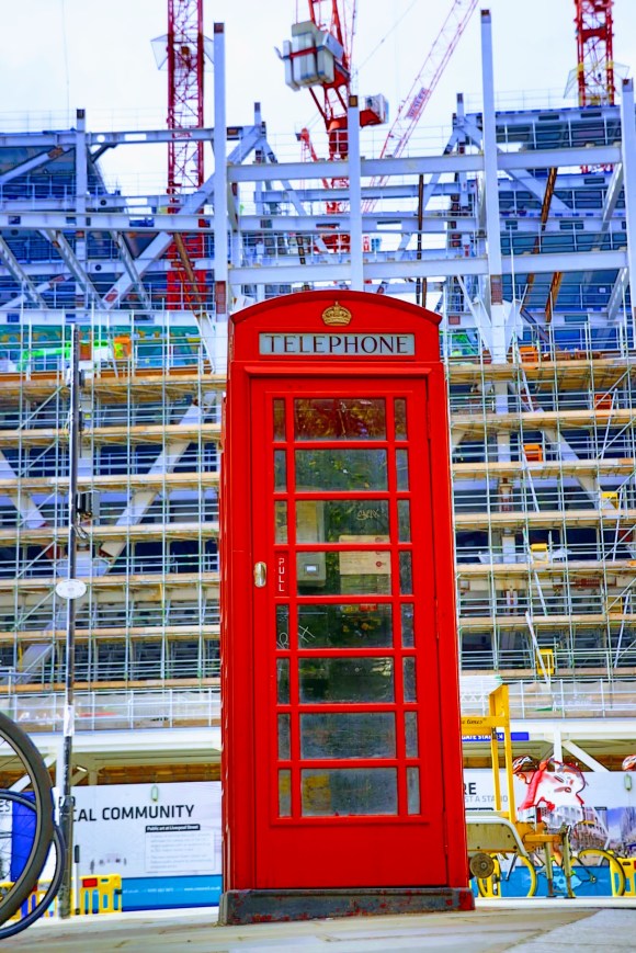 Colour: a ground shot looking up at a red telephone box in the foreground. A scafolding covered building in the background