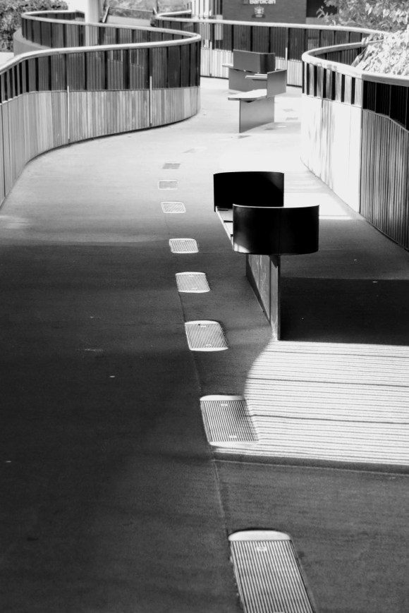 Black and White: an 'S'' curved high level walkway with sides crafted out of two different wood colours. A bench in the foreground with curved ends, and the centre of the floor inlaid with ovoid grills. Sun shining from the left casting shadows